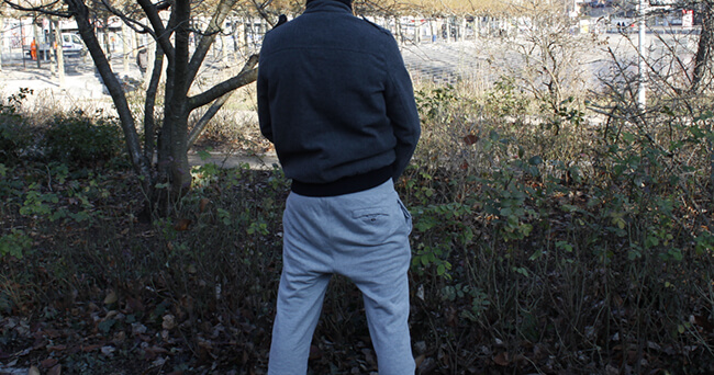 Ein Mann mit grauer Jogginghose und dunkler Jacke steht mit dem Rücken zur Kamera und uriniert in ein Gebüsch am Rand einer öffentlichen Fläche mit Bäumen und Sträuchern.