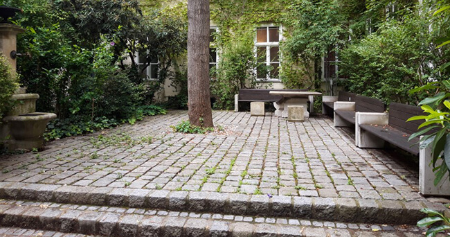 Ein mit Kopfsteinpflaster ausgelegter kleiner Platz ist von Bänken, Bäumen und dichtem Grün umgeben. In der Mitte steht ein Baum, am Rand ein steinerner Tisch mit Sitzhockern.