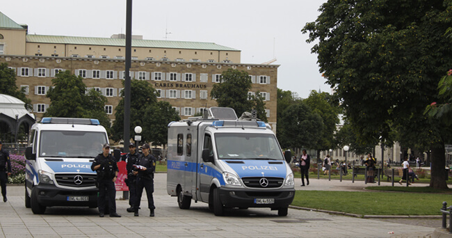 Zwei Polizeiwagen stehen nebeneinander auf einem städtischen Platz. Drei uniformierte Einsatzkräfte unterhalten sich davor. Im Hintergrund sind eine Parkanlage und ein größeres Gebäude zu sehen.