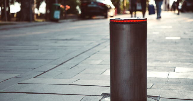 Ein metallischer, versenkbarer Poller mit rotem Leuchtrand steht auf einem gepflasterten Straßenabschnitt. Im Hintergrund sind Fußgänger und parkende Autos sichtbar.