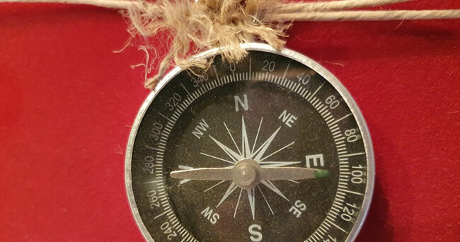 Ein silberner Kompass mit schwarzem Ziffernblatt hängt an einer groben Schnur vor rotem Hintergrund. Symbolisch für Richtung und Orientierung, etwa in einer Präventionsrallye.