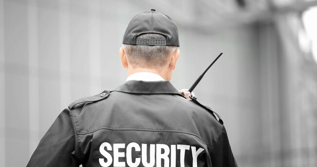 Ein Mann in schwarzer Sicherheitsuniform mit der Aufschrift „Security“ steht mit dem Rücken zur Kamera und hält ein Funkgerät in der rechten Hand.