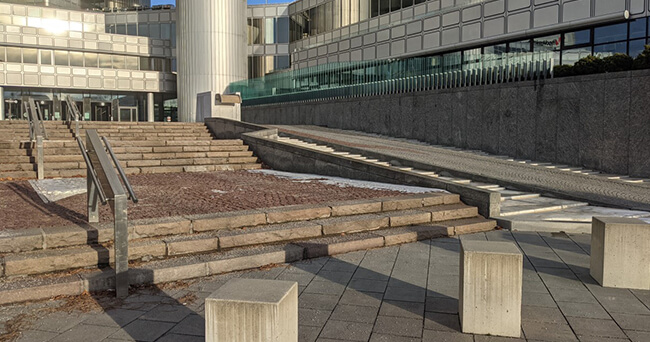 Eine moderne Außenszene mit einer öffentlichen Treppenanlage und daneben liegender Rampe, ergänzt durch Beton-Sitzblöcke im Vordergrund. Die Umgebung ist urban geprägt, mit Glasfassaden und Pflastersteinen. Das Bild steht exemplarisch für einen öffentlich zugänglichen Raum auf Privatgrund (POPS).