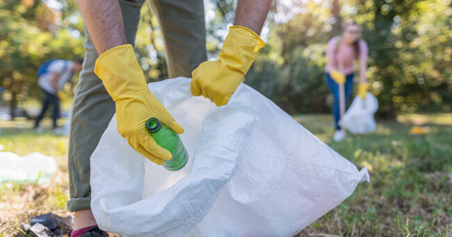 Eine Person mit gelben Gummihandschuhen sammelt eine grüne Plastikflasche in einen großen weißen Müllsack. Im Hintergrund weitere Personen beim Müllsammeln in einem sonnigen Park.