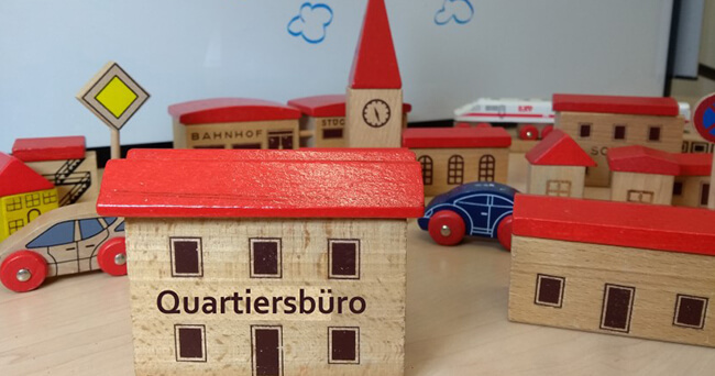 Holzspielzeughäuser mit rotem Dach und der Aufschrift „Quartiersbüro“ im Vordergrund, dahinter weitere Spielzeughäuser, Autos und Straßenschilder auf einem Tisch.