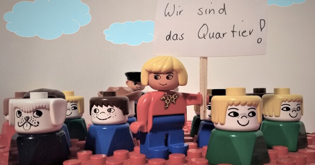 Mehrere bunte Spielfiguren aus Kunststoff stehen auf einer roten Bauplatte, vorne eine blonde Figur mit einem Schild „Wir sind das Quartier!“, im Hintergrund weitere Figuren sowie einfache Wolkenzeichnungen.