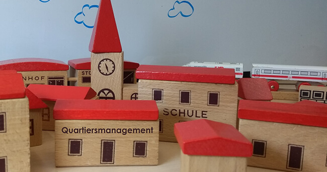 Mehrere Spielzeughäuser mit roten Dächern symbolisieren ein Stadtviertel. Im Vordergrund steht ein Haus mit der Aufschrift „Quartiersmanagement“, daneben weitere Gebäude wie „Schule“ und „Postamt“.