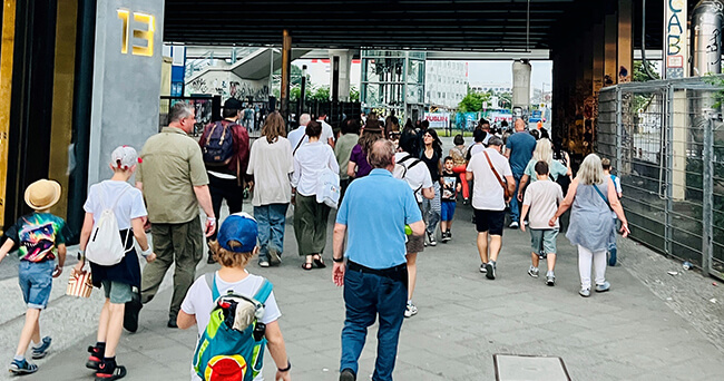 Eine größere Gruppe von Erwachsenen und Kindern geht gemeinsam unter einer Überführung hindurch. Im Vordergrund sind zwei Kinder mit Rucksäcken zu sehen, im Hintergrund weitere Personen auf dem Weg ins Stadtviertel.