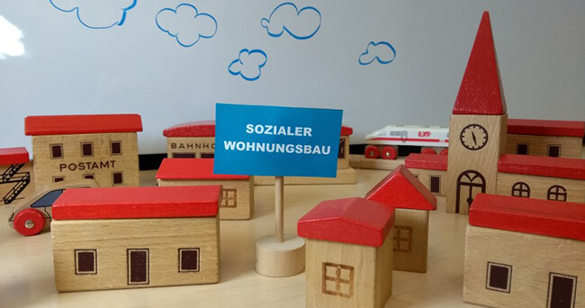 Ein Arrangement aus kleinen Holzmodellhäusern mit roten Dächern, mittig ein Schild mit der Aufschrift „Sozialer Wohnungsbau“. Im Hintergrund gezeichnete blaue Wolken auf einer weißen Tafel.