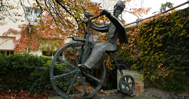 Bronzestatue einer radfahrenden Figur auf einem großen Fahrrad, im Hintergrund ein Gebäude mit herbstlich gefärbtem Efeu bewachsen.