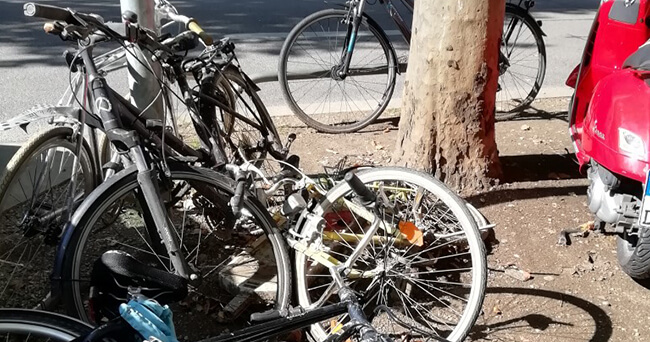 Mehrere beschädigte und verlassene Fahrräder, teils mit verbogenen Rädern, sind an einem Baum und einem Fahrradständer am Straßenrand abgestellt.