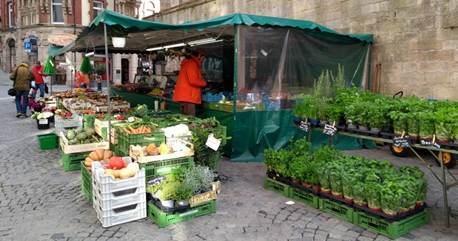 Ein Marktstand mit grünem Pavillon bietet frisches Gemüse, Kräuter und andere Lebensmittel an. Im Vordergrund stehen Kisten mit Kräutertöpfen, im Hintergrund sind weitere Stände und Passanten zu sehen.