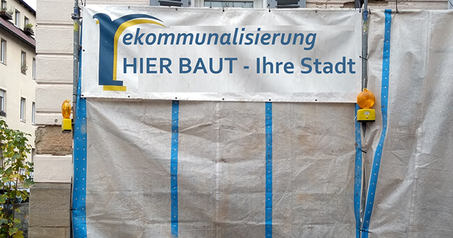 Ein Baustellenschild mit der Aufschrift „Rekommunalisierung – Hier baut Ihre Stadt“, angebracht an einer Hausfassade mit Planen und zwei gelben Warnleuchten.