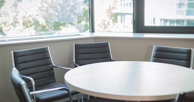 Heller Besprechungsraum mit großem runden Tisch und vier schwarzen Konferenzstühlen vor einem Fenster mit Blick ins Freie.
