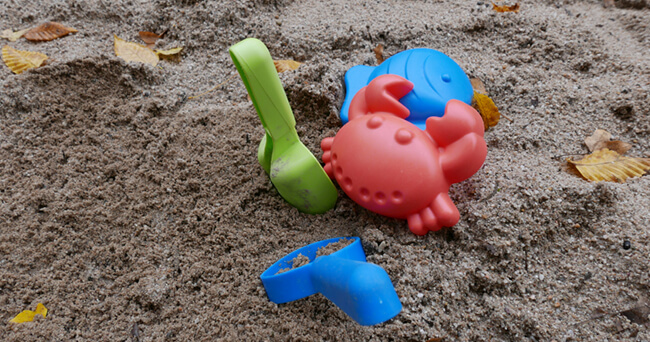 Ein Sandkasten im Freien mit buntem Kinderspielzeug, darunter eine rote Krabbe, eine blaue Form und eine grüne Schaufel, umgeben von Sand und einzelnen Blättern.