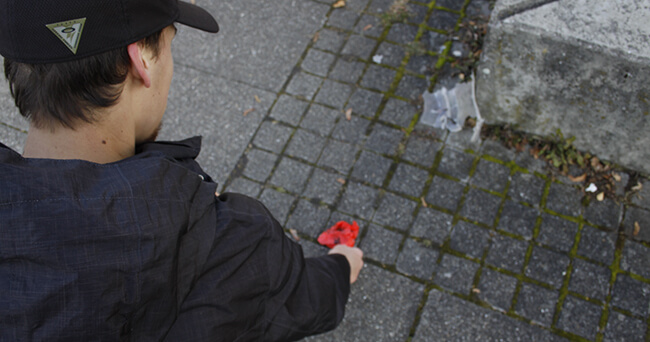 Eine Person in schwarzer Jacke und schwarzer Mütze bückt sich, um auf einer gepflasterten Fläche ein Stück roten Müll aufzuheben.