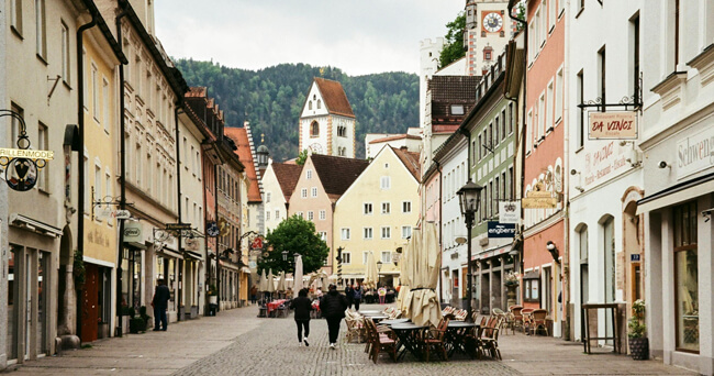 Blick in eine autofreie Innenstadt mit Menschen auf der Straße, Gastronomie im Freien, alten Häuserfassaden und einer Kirche im Hintergrund. Die Straße wird gemeinsam von Fußgängerinnen und Fußgängern sowie Radfahrenden genutzt.