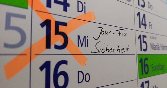 Nahaufnahme eines Wandkalenders mit orange markiertem Datum und handschriftlichem Eintrag „Jour-Fix Sicherheit“.