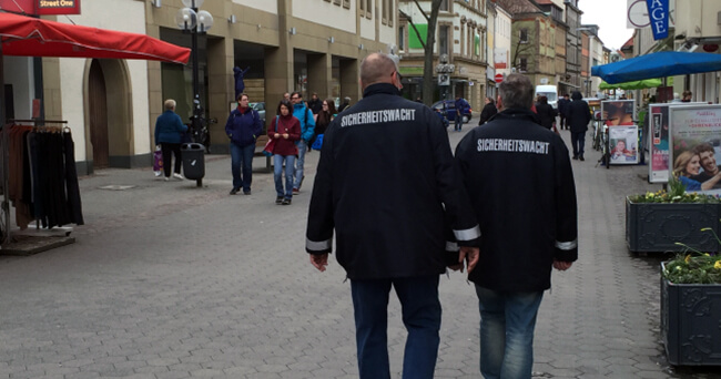 Zwei Sicherheitswacht-Mitarbeiter in Uniform gehen durch eine belebte Fußgängerzone in einer Innenstadt.