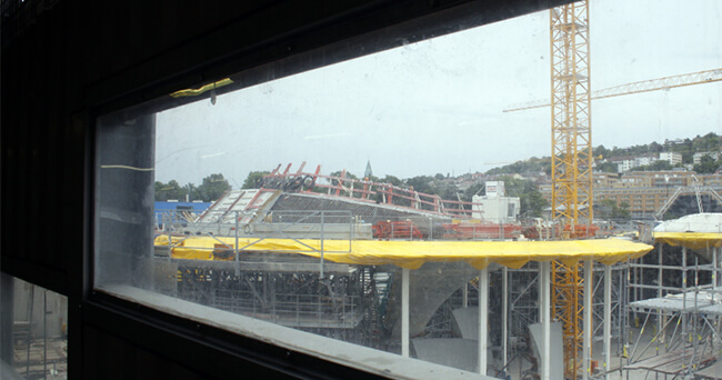 Foto einer Baustelle, aufgenommen durch ein rechteckiges Sichtfenster in einer Absperrung. Im Hintergrund sind Baukräne, Gerüste und ein teilweise überdachtes Gebäude zu sehen.