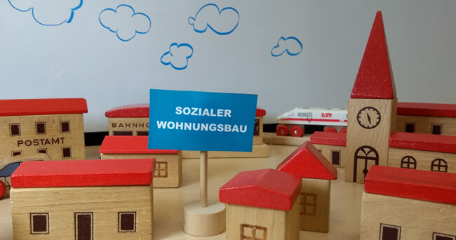 Mehrere Holzbausteine in Form von Häusern mit roten Dächern, im Vordergrund ein Schild mit der Aufschrift „Sozialer Wohnungsbau“, im Hintergrund eine Spielzeugkirche, ein Postamt und weitere Gebäude.
