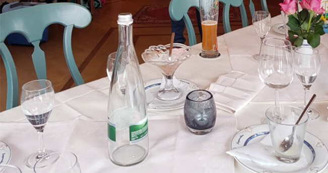 Ein festlich gedeckter Restauranttisch mit mehreren Gläsern, Tellern und einer leeren Wasserflasche, aufgenommen nach dem Ende einer Veranstaltung.
