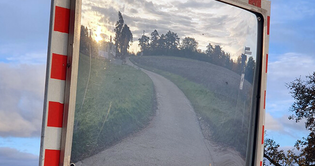 Ein Verkehrsspiegel mit rot-weißer Umrandung zeigt den Blick auf einen schmalen, geschwungenen Weg in ländlicher Umgebung bei Sonnenuntergang.