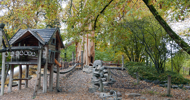 Ein großer öffentlicher Spielplatz in einem Park mit Klettergerüsten, Holzhäuschen, Rutschen und Netzen, umgeben von herbstlich gefärbten Bäumen.