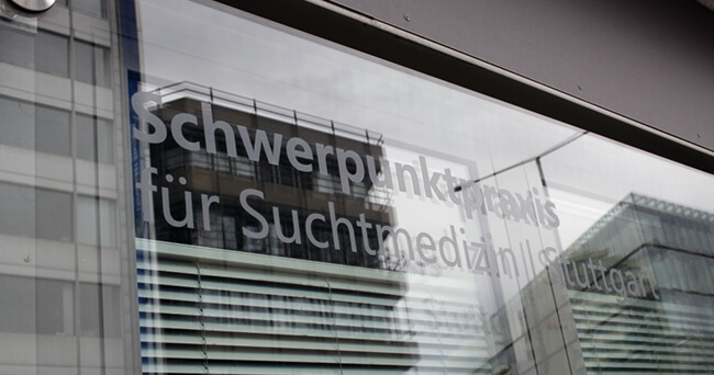 Blick auf die Glasfront einer Substitutionspraxis mit Schriftzug „Schwerpunktpraxis für Suchtmedizin“, in der sich Gebäude und Himmel spiegeln.