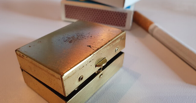 Ein goldfarbener, rechteckiger Taschenaschenbecher liegt auf einem weißen Tisch, dahinter eine Streichholzschachtel und eine Zigarette.