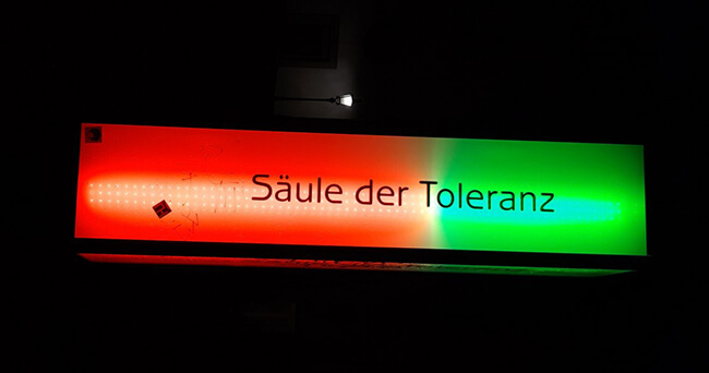 Beleuchtetes Schild bei Nacht mit der Aufschrift „Säule der Toleranz“, links rot, rechts grün beleuchtet, vor dunklem Hintergrund.