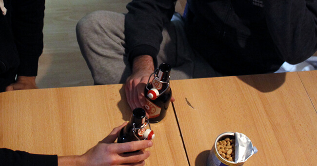 Alt-Text Zwei Personen sitzen an einem Holztisch, jede hält eine Bierflasche in der Hand; daneben steht eine geöffnete Dose mit Erdnüssen.