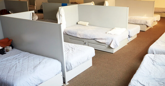 Mehrere einzelne Betten mit weißer Bettwäsche, durch Sichtschutzwände voneinander getrennt, stehen in einem beheizten Raum für obdachlose Menschen bereit.