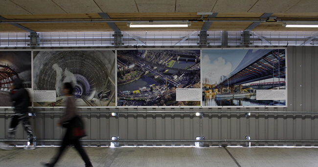 Blick durch eine moderne Unterführung mit mehreren großformatigen Ausstellungstafeln an der Wand, zwei unscharfe Passanten gehen daran vorbei.