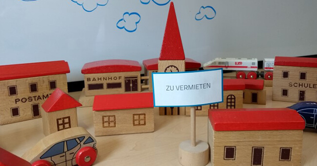 Mehrere kleine Holzmodelle von Häusern, Autos und einer Kirche mit rotem Dach, davor ein Schild mit der Aufschrift „Zu vermieten“.