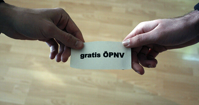 Zwei Personen halten gemeinsam ein kleines Schild mit der Aufschrift „gratis ÖPNV“ vor einem hellen Hintergrund.