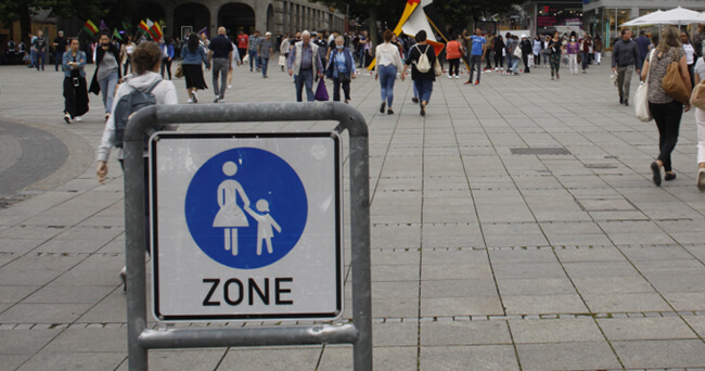 Menschen laufen über einen weiten, gepflasterten Platz mit Zonenbeschilderung im Vordergrund, keine Autos sichtbar, im Hintergrund eine belebte Innenstadt.