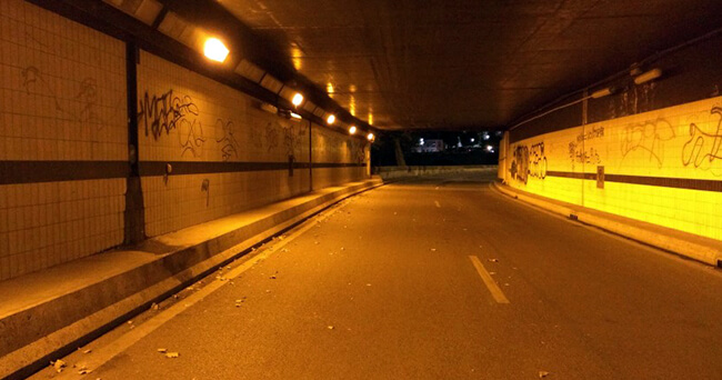 Leere, gelb beleuchtete Straßenunterführung mit Graffiti an den Wänden und leichtem Müll auf der Fahrbahn.