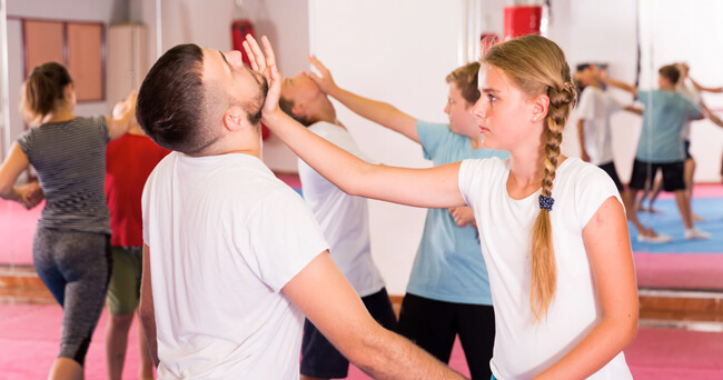 Mehrere Jugendliche trainieren Selbstverteidigungstechniken in einer Turnhalle, im Vordergrund wehrt ein Mädchen einen simulierten Schlag eines jungen Mannes ab.