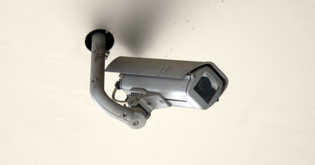 An einer weißen Decke installierte silberfarbene Überwachungskamera mit Wandhalterung, leicht seitlich nach unten ausgerichtet.