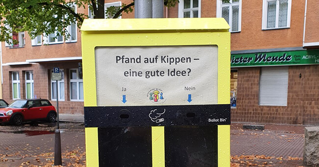 Gelber Abstimmungsbehälter für Zigarettenkippen mit der Frage „Pfand auf Kippen – eine gute Idee?“ und den Antwortmöglichkeiten „Ja“ und „Nein“, aufgestellt an einer Straßenlaterne in einer Wohngegend.
