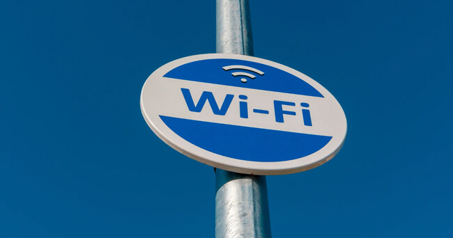 Ein rundes, blau-weißes Schild mit der Aufschrift „Wi-Fi“ ist an einem Mast befestigt. Im oberen Teil des Schildes befindet sich ein WLAN-Symbol, im Hintergrund ist ein klarer blauer Himmel zu sehen.