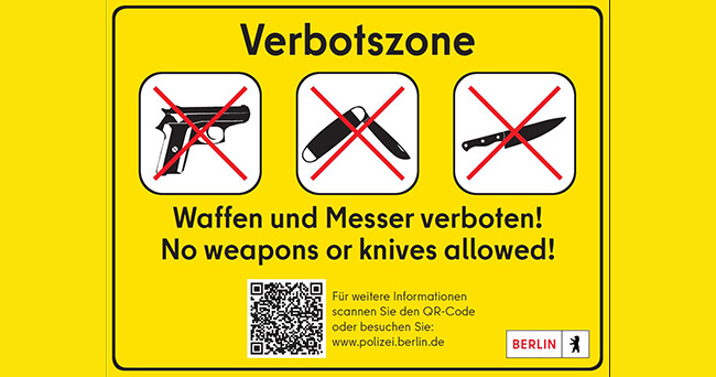 Gelbes Hinweisschild mit Piktogrammen von durchgestrichenen Waffen und Messern, Text „Waffen und Messer verboten! No weapons or knives allowed!“ sowie QR-Code und Logo der Polizei Berlin.