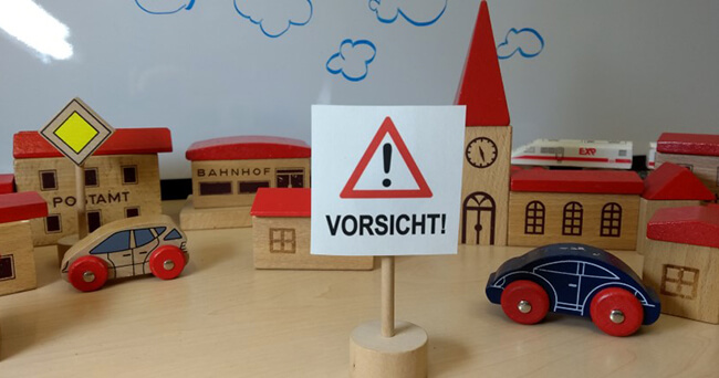 Ein Holzmodell einer kleinen Stadt mit Postamt, Bahnhof, Kirche, Schule und Autos. Im Vordergrund steht ein Schild mit einem roten Warndreieck und dem Schriftzug „Vorsicht!“.