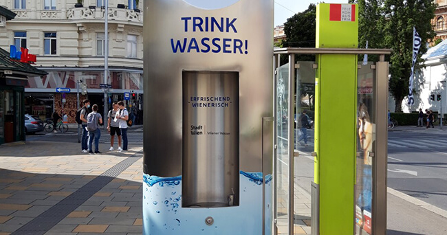 Edelstahl-Wasserspender mit der Aufschrift „Trink Wasser!“ steht an einer belebten Straßenecke, im Hintergrund sind Passanten und Geschäfte zu sehen.