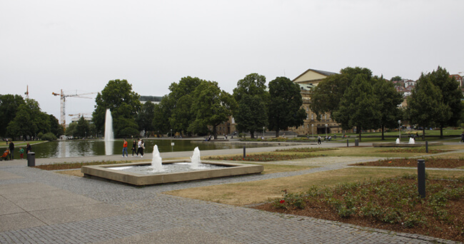 Blick auf einen öffentlichen Platz mit mehreren Wasserspielen und Springbrunnen, umgeben von Bäumen und Gehwegen, im Hintergrund ein großes Gebäude und Baukräne.