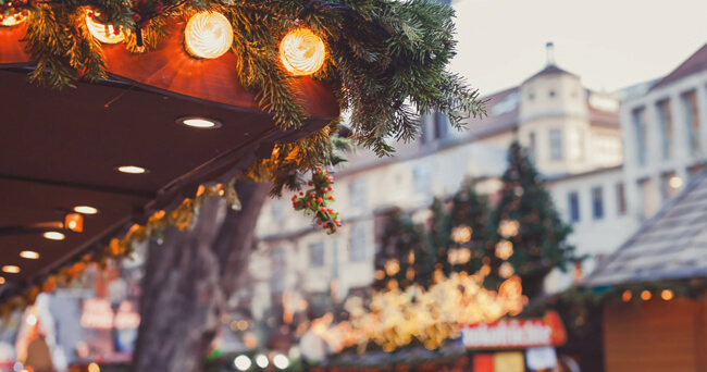 Nahaufnahme eines weihnachtlich dekorierten Marktstandes mit Lichterkette und Tannenzweigen, im Hintergrund unscharf beleuchtete Gebäude und weitere Stände eines Weihnachtsmarkts.