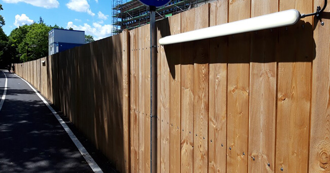 Langer, stabiler Bauzaun aus Holz entlang einer Baustelle mit montierter Leuchte und angrenzendem Fuß- und Radweg bei sonnigem Wetter.