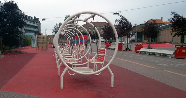 Runde Metall-Sitzkonstruktionen auf einem Platz mit rotem Bodenbelag, dahinter weitere Sitzgelegenheiten und städtische Umgebung.