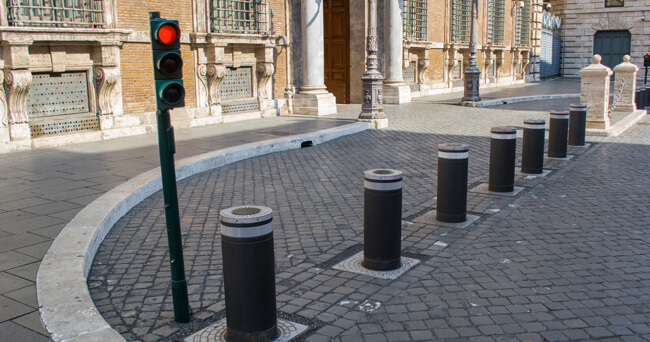 Reihe fest installierter, zylindrischer Poller vor einer Straßeneinfahrt, daneben eine Ampel mit rotem Signal, um unbefugte Fahrzeugzufahrt zu verhindern.